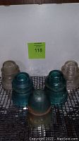 View of seven Hemingray glass insulators on mesh shelf, including five clear and two teal blue ones, showing wear and some chips.