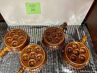 Four round brown stoneware escargot/snail dishes arranged on a black mesh surface. Each dish has six wells and a handle.
