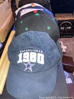 Photo shows a Dallas Cowboys baseball hat embroidered with 'ESTABLISHED 1980' and the team's star logo, placed on top of a stack of hats.