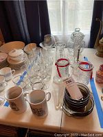 Full view of table with glassware and ceramics including stemware, tumblers, ceramic mugs, cut glass lidded jar, boot mugs and custard cups