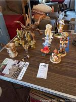Photo showing seven Goebel Hummel girl figurines with pastoral and playful themes, placed on a wooden surface alongside a natural wood plant stand and two candlesticks.