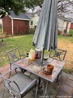 Full view of slate top square patio table with umbrella and three metal frame chairs with cushions set on patio bricks in backyard environment with shed behind.