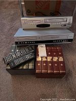 Photo showing Emerson and Philips combo players stacked, with VHS tape cases and remotes in front on carpeted floor.