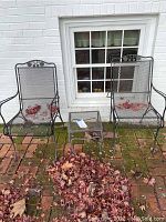 Lot showing two black metal mesh patio chairs and one small metal mesh end table on an outdoor brick patio with leaves around them.
