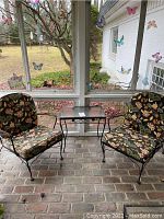 Full view of the two metal patio chairs with floral patterned cushions and the matching glass-topped accent table situated on a screened porch with brick flooring.