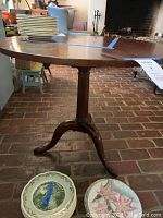 Side view of the pedestal table showing the tripod base with curved legs and mahogany finish on a brick floor with plateware on top.