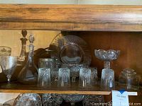 Full view of assorted crystal glassware on wooden shelf including glasses, decanter, bowl, candy dish and platter