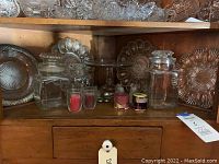 Wood cabinet shelf with glassware displayed including three sizes of glass cookie jars, glass pedestal cake platter, some jars with lids, and decorative crystal platters underneath.