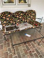 Patio sofa with black metal frame and floral patterned cushions alongside a rectangular glass top coffee table on a brick floor.