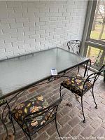 Photo showing angled view of glass top metal patio table with two metal chairs with floral seat cushions on brick floor near white brick wall and windowed porch area.