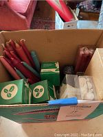 Box containing multiple red taper candles along with some green boxed candles featuring holiday holly graphics, plus some other shaped candles and candleholders.