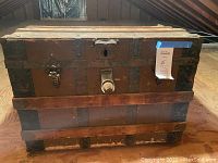 Front view of vintage wooden trunk with metal bands and fittings, showing lock mechanism and seller tag.