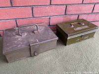 Two old metal strong boxes placed side by side against a brick wall, showing the relative size and condition.