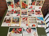 Photo showing the full set of 15 vintage Coca-Cola ads laid out on a green surface. Each ad is a full-color illustration from old National Geographic magazines, prominently featuring Coca-Cola branding and nostalgic scenes.