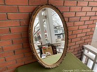 Front view of the oval mirror showing the reflective surface and decorative gold-tone frame against a brick wall.