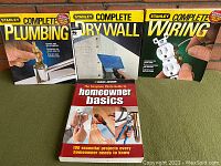 Four DIY home repair books laid flat on a green surface in front of a brick wall, showing the front covers for identification and condition.