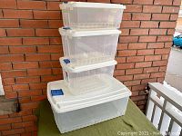 Set of four clear Rubbermaid storage bins stacked outside against a brick wall. Shows four bins with white lids and blue latches.