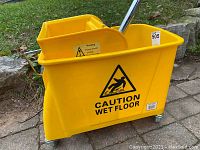 Yellow plastic mop bucket with built-in wringer and metal handle. Marked with caution wet floor symbol. Bucket has metal wheel braces with caster wheels.
