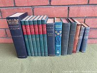 Full view of 13 old decorative books arranged upright against a brick wall, showing varied sizes and colors.