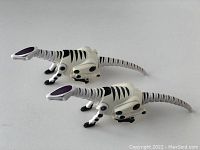 Two white robotic reptile toys with black stripes and purple accents on heads, side view
