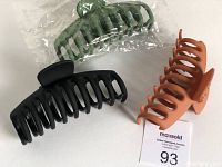 Photo showing three large plastic hair claw clips in black, matte orange, and dark green. Two clips are loose, one still bagged.