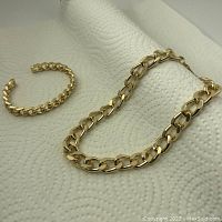 Goldtone chain necklace and matching bracelet set photographed on a white textured background, showing the linked design and polished finish.
