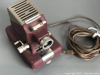 Angled side view showing textured burgundy metal casing, ventilation slots, silver lens mount, and attached long electrical cord.