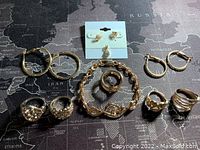 Full view of assorted gold tone jewelry laid out on a black and white world map background. Includes rings, various styles of hoops and stud earrings, a bracelet, and a pendant earring or pendant.