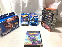 Front view of multiple Hot Wheels toy cars in original packaging, organizing tin box, and World Race DVD spread on a light-colored surface with a light backdrop.