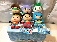 Front view of 6 plush toys showing the characters Green Lantern, Catwoman, Joker, Wonder Woman, Superman, and Batman on a silver background, with tags and box beneath.
