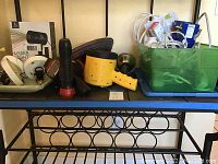 Photo showing various items on a blue shelf including ceiling light fixture parts, a yellow LED laser light projector, four black flashlights, multiple extension cords, power strips, light bulbs, and batteries stored in a green bin.