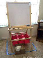 Front view of the easel showing dry erase board with markers, eraser, and cleaner on the tray below, and red fabric storage bins underneath with a box of coloring books on the floor.