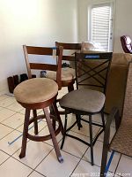 Three bar stools grouped together showing two wooden stools with beige suede seats and one black metal stool with grey fabric seat.
