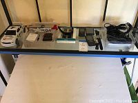 Top shelf displaying white RC car, silver P-51 Mustang RC airplane model on box with stand and manual, black Metro PCS flip phones, unbranded cell phone, black quartz wrist watches with bands, fitness wrist device, and various charging cables.