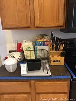 Photo showing metal dish rack, plastic cutting board, various kitchen storage items, paper products, and a red cup on countertop under kitchen cabinets.