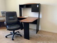 View of black padded office chair with wheels and L-shape desk with wood-tone top and black sides and hutch.