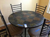 Round multi-colored tile top dining table with 4 cushioned metal back chairs on a rectangular patterned rug in a dining room.