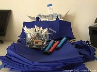 Stack of folded blue fabric tote bags with clear container holding assorted pens and capped markers on top, with LED clip-on flashlights in front.