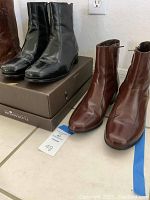 Two pairs of Florsheim men's dress boots placed on floor, one black pair on box and one brown pair beside it.