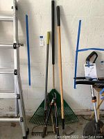 Photo showing garden rake, metal rake, garden hoe leaning against a wall next to a ladder in a garage.