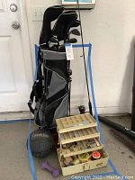 Golf bag filled with assorted golf clubs with head covers, fishing tackle box filled with accessories in front, and other items around on the floor.