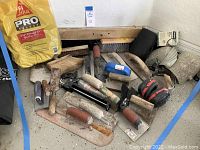 Photo showing a variety of hand tools including trowels, scrapers, a black caulking gun, brooms, and gloves all exhibiting usage wear and dirt.