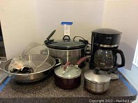 Full lot front view showing the Crock-Pot, coffee maker, pots with lids, electric skillet without lid, and metal strainer, all placed on a kitchen counter.