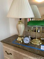 Photo showing cream ceramic table lamp with brass base and cream shade, green glass banker-style desk lamp, and silver-tone alarm clock