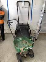 Tornado 16 inch manual reel push mower with grass catcher bag and Scotts Turf Builder EdgeGuard Mini fertilizer spreader together in garage.