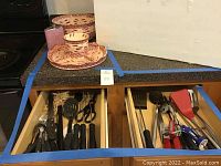 Overview of kitchen drawers open showing assorted kitchen utensils with black handles, and surface showing ceramic serving items and flask
