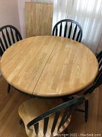 Photo of round table with natural wood top and dark painted legs, four matching chairs around it, and one leaf leaning against the wall