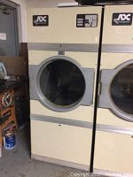 Front view of large commercial gas dryer showing circular transparent door and beige-gray color scheme