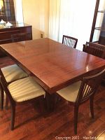 Photo shows rectangular wood dining table with six matching chairs around it. Table finish is polished and some wear visible on surface. Chairs have vertical slat backs and light upholstered seats.