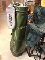 Green golf bag containing left hand golf clubs, showing wear, next to folded camp chairs.